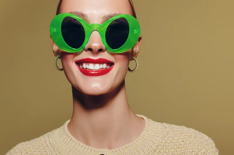 Smiling woman with bright red lipstick wearing large green sunglasses and hoop earrings. Close-up portrait on beige background. Concept of fashion, style, and summer accessories.