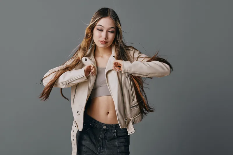 Young woman with long hair wearing leather jacket and crop top posing against gray background. Fashion model showing casual style with confident expression and natural makeup.
