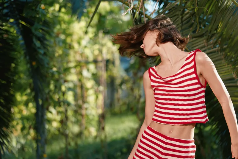A cheerful woman wearing a red and white striped crop top and skirt walks through a sunny garden, hair in motion, capturing a playful, carefree summer mood.