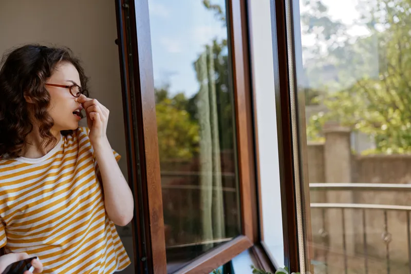 Woman in striped shirt stands by an open window, enjoying natural daylight, a candid home moment that conveys relaxed lifestyle and contemplation, and a hint of morning air fills the scene creating a