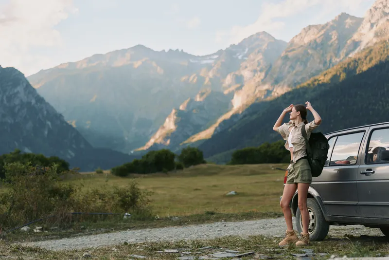 Young woman with backpack stands near a car in a mountain valley enjoying the scenic view on a clear day during a hiking trip.
