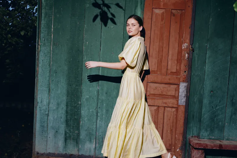 A woman in a flowing yellow dress walks past a green wall and wooden door, captured in an outdoor fashion shot with soft evening light and a candid street style vibe