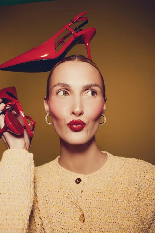 Young woman with red lipstick holds red high heel shoes near her head, wearing yellow sweater and gold hoop earrings. Studio portrait with warm lighting and playful expression.