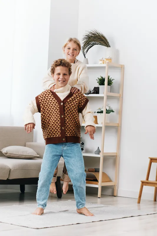 Two children playfully pose indoors, a girl perched on a smiling boy's shoulders, mid action and laughter, in a bright cozy living space with soft light.