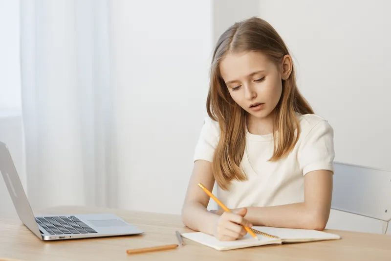 Thoughtful young girl writing in a notebook at a desk, captured in a bright and minimalistic workspace with a laptop, reflecting focus and concentration on her schoolwork