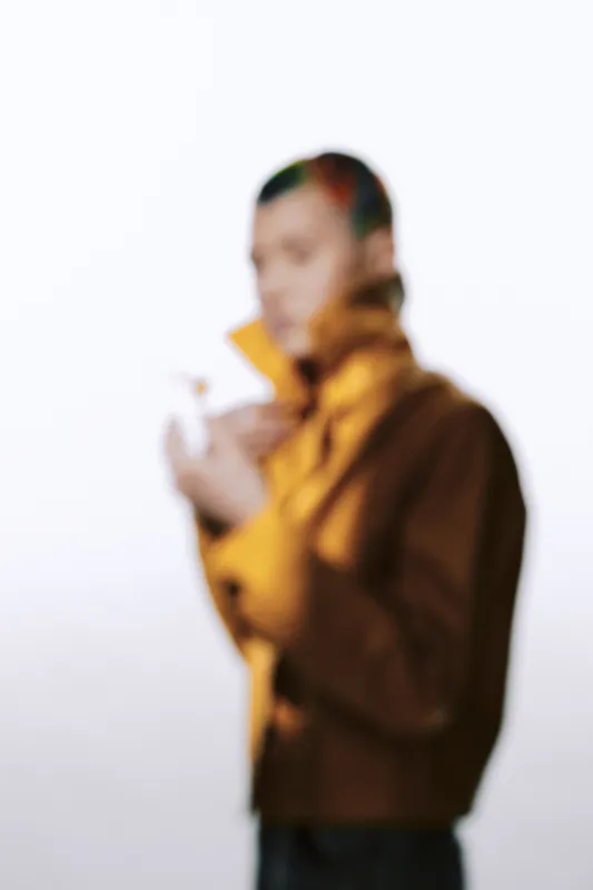 Blurry woman wearing a brown jacket stands against a light background, creating a soft and mysterious atmosphere with muted colors and casual style.
