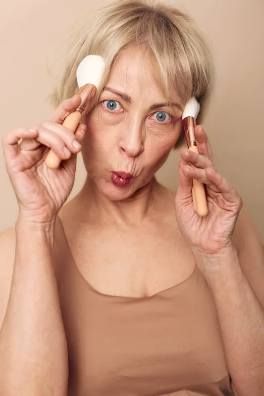 Mature woman with blue eyes holding two makeup brushes near face making playful expression on beige background. Concept of beauty, skincare, cosmetics, and joyful self-care for older adults.