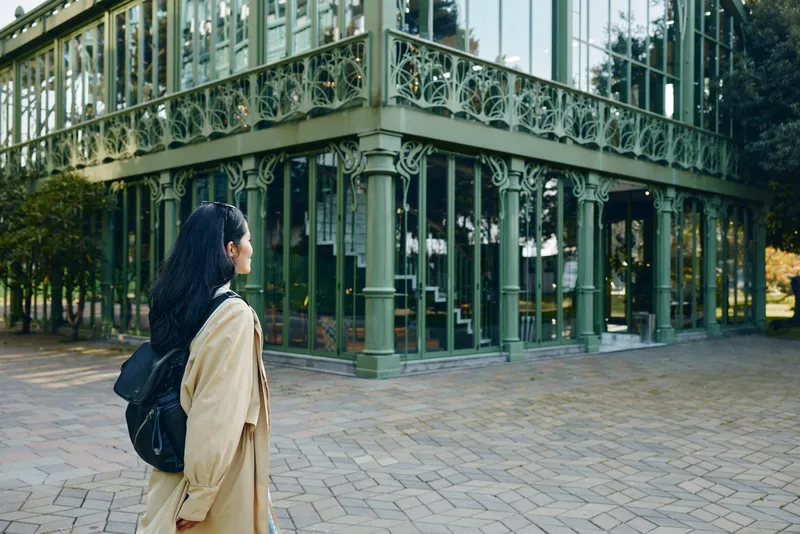 Woman with backpack gazing at a glass conservatory building, outdoor architectural setting, reflective surfaces, calm exploration mood, urban garden vibe and morning light