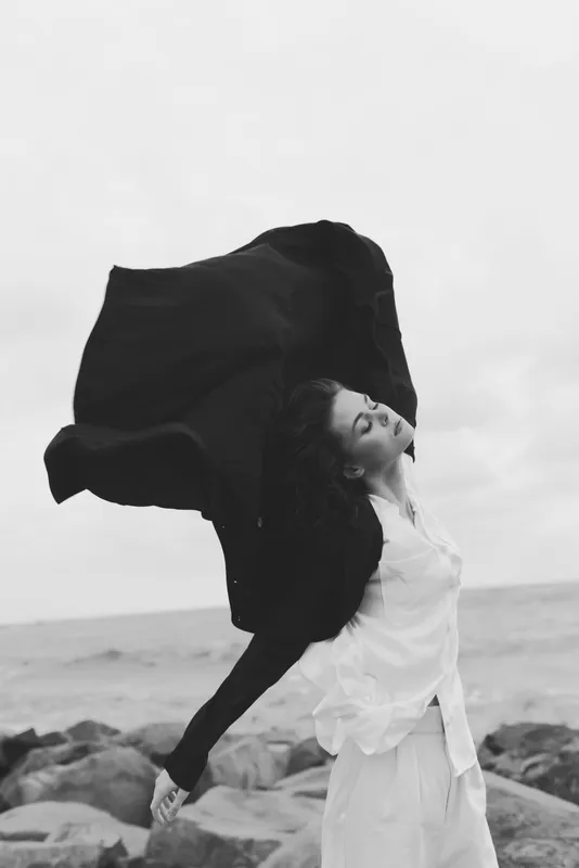 Wind sweeps a cape over a white dress as a woman stands on a rocky shore, sea and sky behind, capturing motion, drama, and monochrome beauty