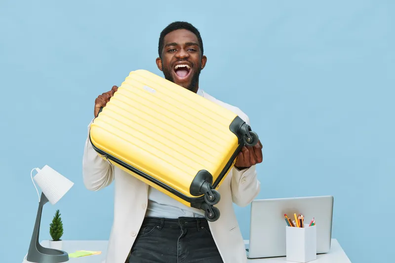 Smiling excited man holding yellow suitcase in office space with blue background, work desk with laptop, stationery, and lamp, expressing happiness and travel enthusiasm, casual attire, lifestyle conc