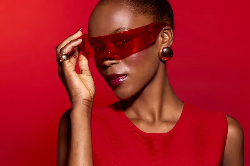 Elegant dark skinned African woman wearing red sleeveless dress and red sunglasses poses against a red background. Concept for fashion, style, beauty, and confidence.