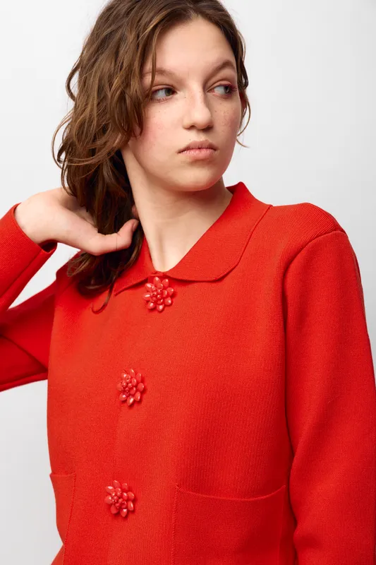 Young woman in red textured sweater with floral buttons looks sideways with thoughtful expression. Studio portrait on white background. Concept of fashion, style, and casual wear.