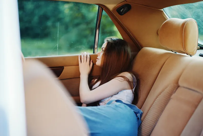 Thoughtful young woman with long hair sitting in a car, gazing out the window, captured in a serene moment with warm tones and a natural background.
