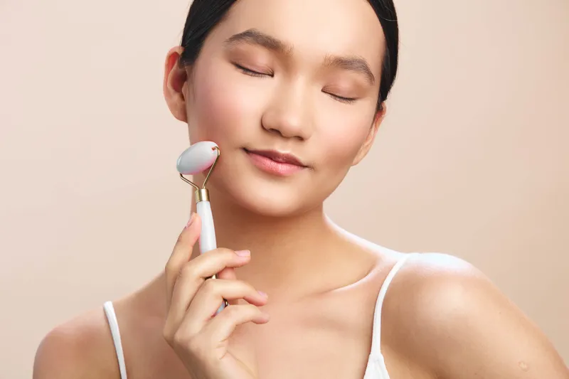 Asian woman enjoys a calming skincare moment, smiling softly while using a facial roller. The light backdrop highlights gentleness, wellness, and a peaceful selfcare routine.