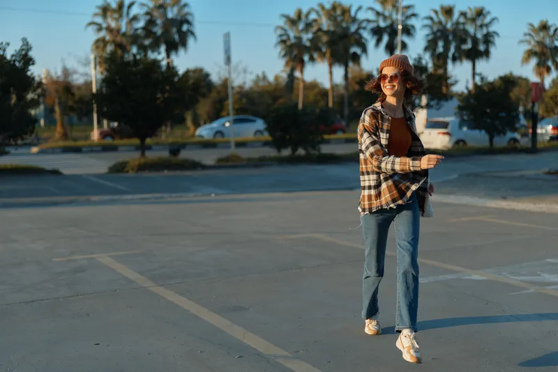 Woman walking with coffee and sunglasses, wearing beanie in a parking lot at golden hour, candid urban portrait capturing authenticity, mindful living and emotional storytelling vibe.