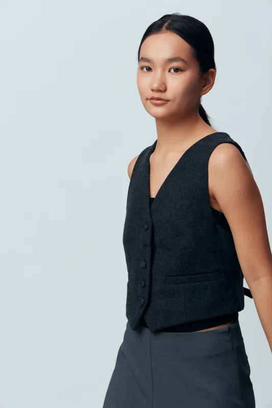 Young woman portrait with calm expression wearing black vest and dark trousers standing against plain light grey background. Asian female model posing with natural makeup and tidy hair in studio