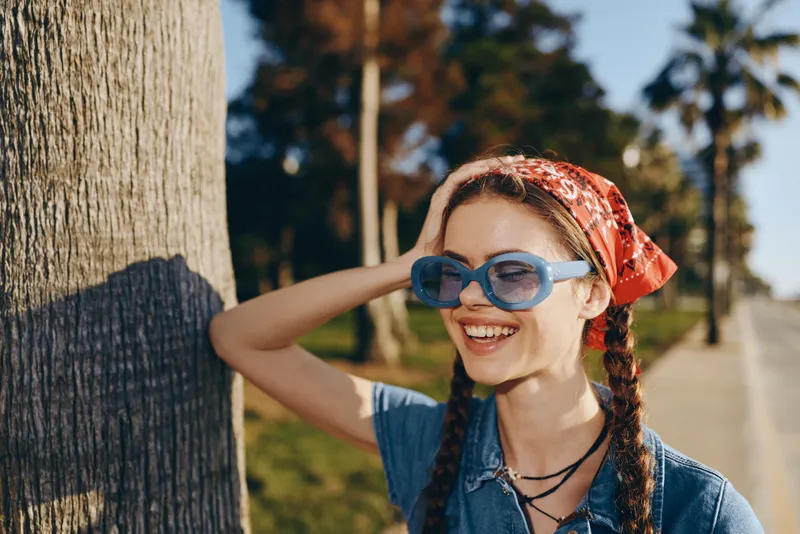Lifestyle woman in modern boho-western denim street style smiling outdoors with braided hair, blue sunglasses, and a red headscarf in film color tone.