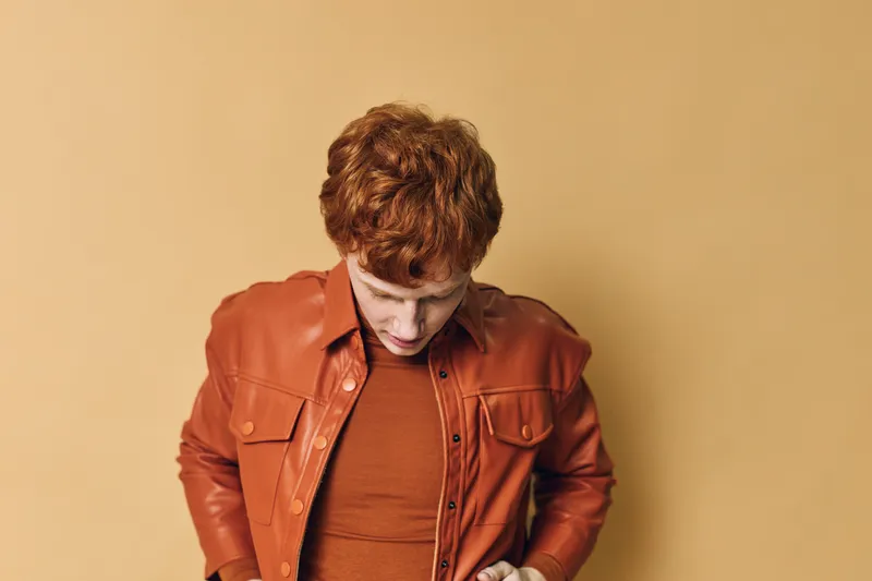 Young man with red hair wearing red shirt and leather jacket stands against beige background looking down with thoughtful expression. Casual fashion and style portrait.