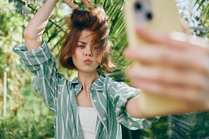 Young woman takes a selfie outdoors in a sunny tropical garden, wind tousling hair, wearing a casual striped shirt, with vibrant greenery in the background.
