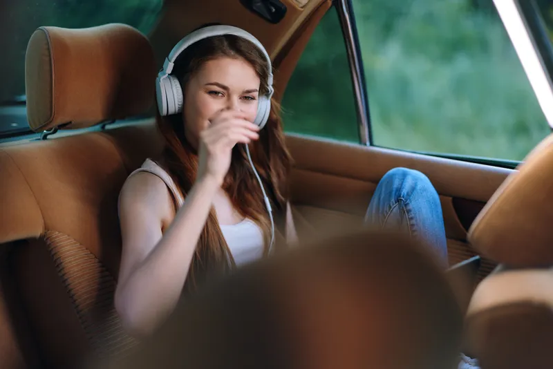 Young woman enjoying music in a car, expressing relaxation and joy, wearing casual clothing, with a blurred green background and warm brown tones.