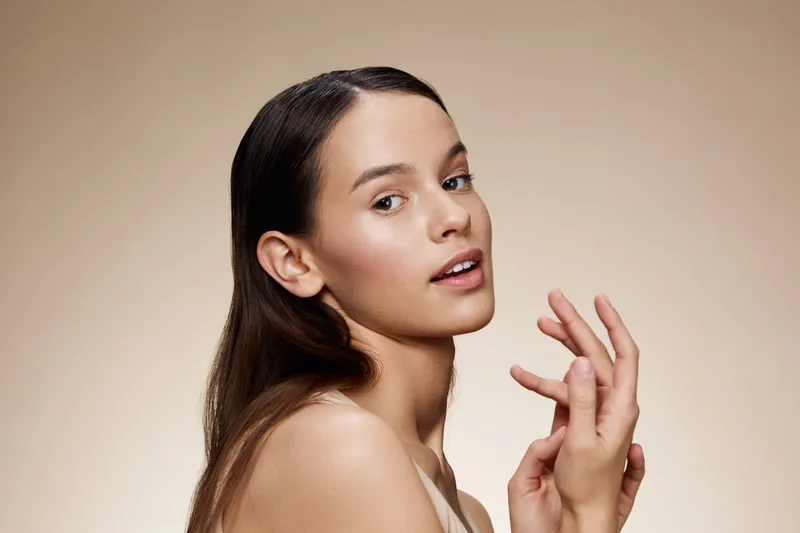 Beautiful young woman with smooth skin, natural makeup, and healthy hair posing against a soft, neutral background, embodying confidence and elegance