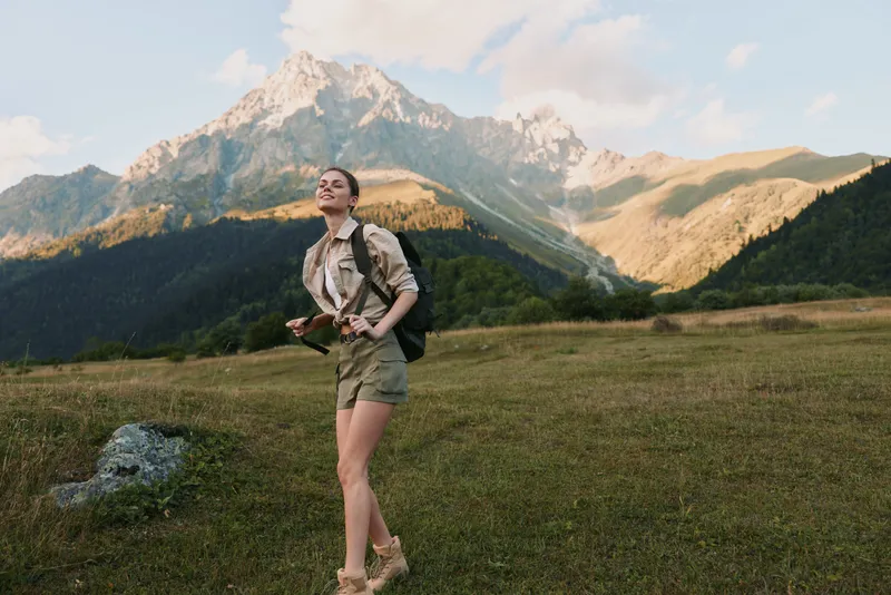 Young woman with a backpack enjoys hiking in a mountain meadow while holding a map and smiling under a clear sky during a sunny day.