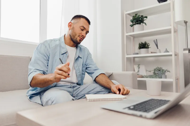Thoughtful man writing in notebook while sitting on sofa, inspired by bright natural light. Ideal for showcasing creativity and productivity at home.