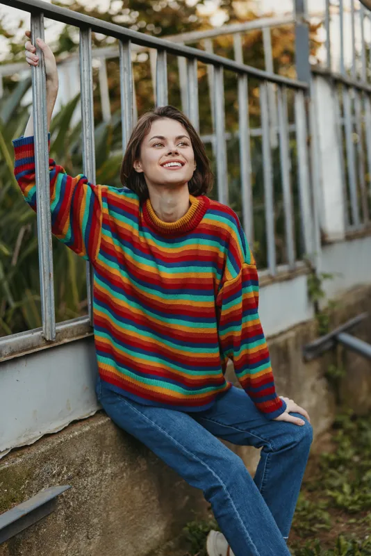 Woman in a colorful striped sweater smiling and leaning on a metal railing, casual outfit with jeans in an outdoor urban garden. Relaxed lifestyle portrait with autumn travel vibe.