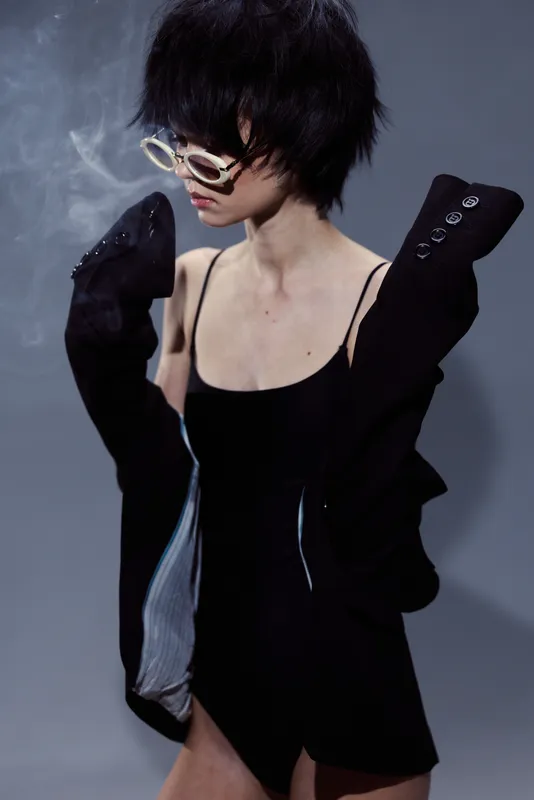 stylish woman with glasses wearing black clothing in moody lighting with smoky background and dramatic pose