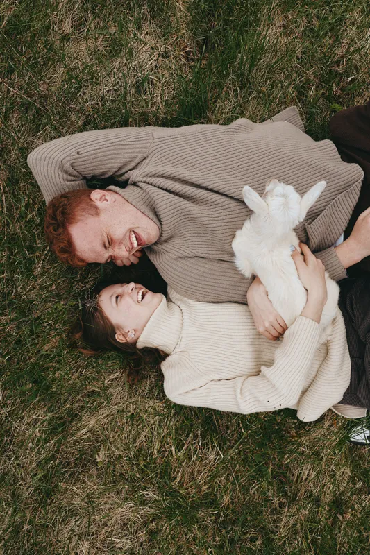 Lifestyle couple outdoors on grass, cuddling a goat kid, warm smiles, cozy sweaters, natural colors, peaceful moment, tender affection, sunlit scene, relaxed pose, candid portrait, intimate connection