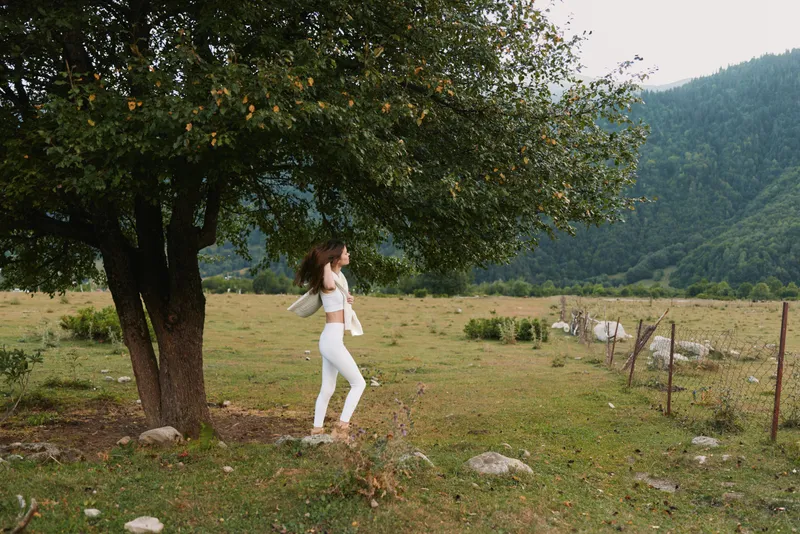 Young woman in white clothes walks near a large tree in a green meadow surrounded by mountains on a cloudy day.