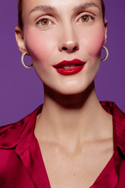 Close-up portrait of a woman with red lipstick and hoop earrings wearing a red blouse on purple background. Concept of beauty, fashion, and makeup style.