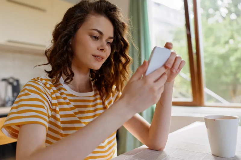 Casual woman in a striped t shirt sits at a sunlit table, focused on a smartphone. A warm, relaxed home scene highlighting connection, comfort, and everyday technology use.