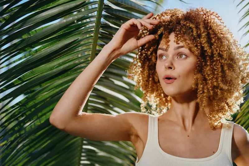 Curious woman with curly hair wearing white tank top standing among green palm leaves in bright natural sunlight, showcasing summer vibe and tropical nature atmosphere.