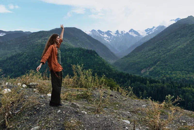 Woman standing on a rocky ledge in a dramatic mountainous landscape with arms raised, enjoying a vast valley, blue sky, and fresh alpine air
