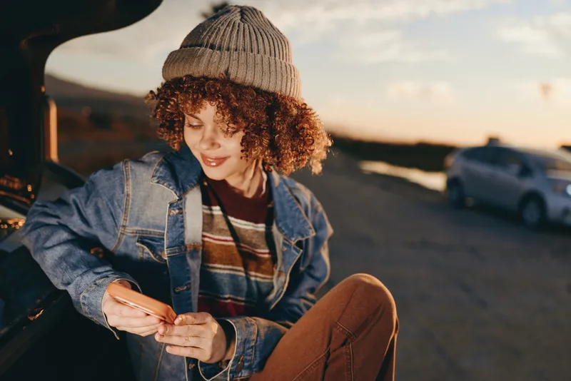Woman lifestyle outdoors at sunset, sitting by a car, wearing denim jacket and knit beanie, curly hair, looking down at phone, warm light, casual mood, urban street background
