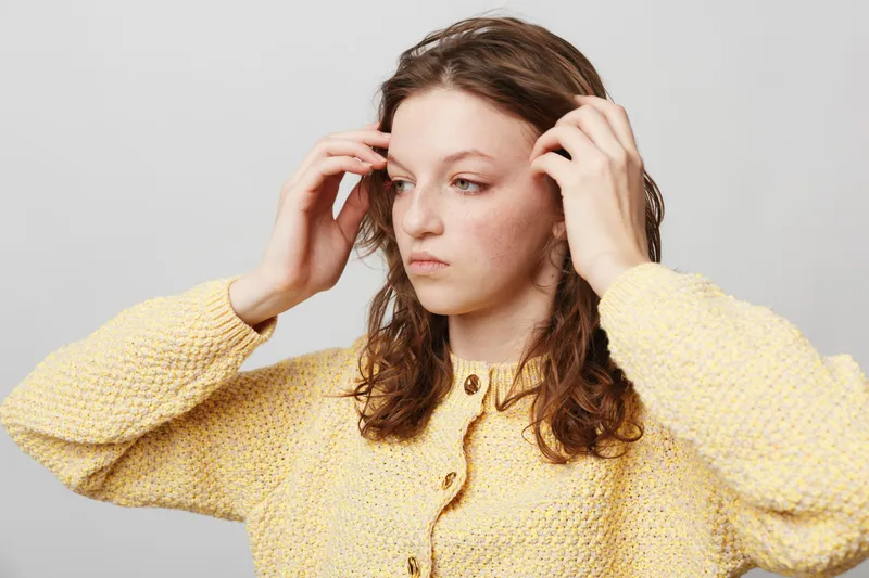 Young woman with wet hair wearing yellow knitted sweater adjusts hair with hands. Neutral background and natural expression. Concept of casual style and personal care.