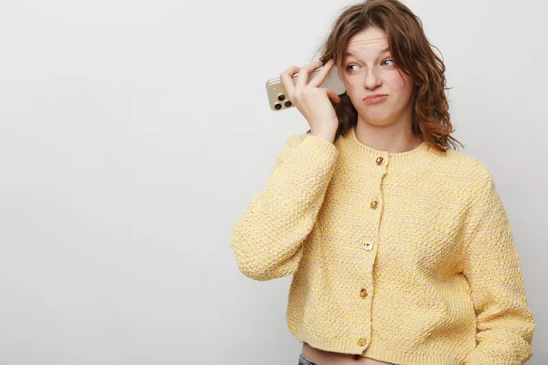 Young woman in yellow sweater holds smartphone near head with confused expression on white background. Concept for doubt, uncertainty, and indecision in communication or technology.