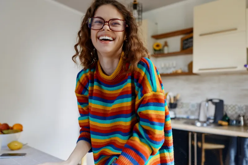 Young woman with curly hair wearing a colorful striped sweater laughs in a bright kitchen, conveying a cheerful home lifestyle moment