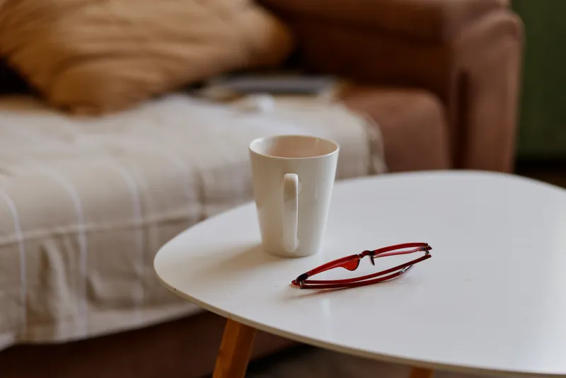 Cozy indoor living room scene with a white mug and red glasses on a round coffee table, a soft couch in the background, and a relaxed cat nearby while warm natural light fills the space