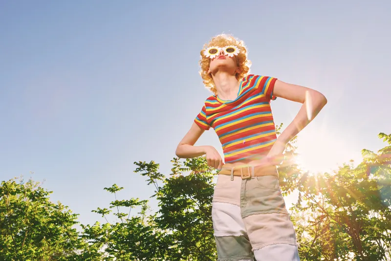 A retro fashion inspired woman outdoors in sunny light, wearing a striped shirt and sunglasses, exuding carefree vibe and vibrant style in a bright outdoor setting.