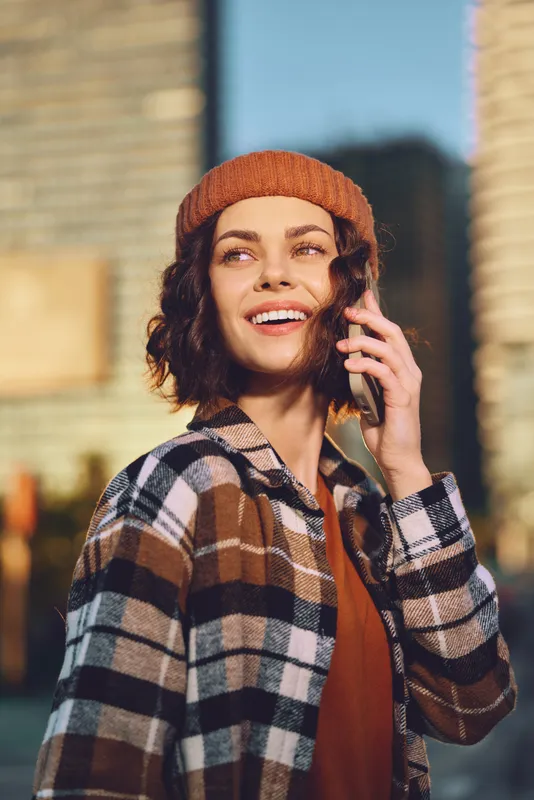 Woman on phone smiles in city portrait wearing beanie and plaid jacket, outdoor sunlight casting golden hour glow. Authenticity and candid lifestyle captured with mindful living vibe.