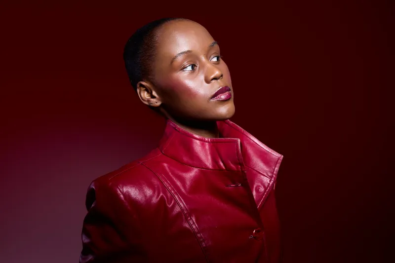 Confident African woman with dark skin wearing a red leather jacket poses against a deep red background. Studio portrait with dramatic lighting highlights facial features and expression.