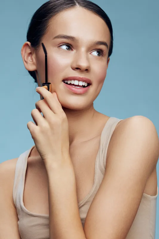Happy young woman showcasing beauty product with a smile against a light blue background, demonstrating confidence and skincare routine