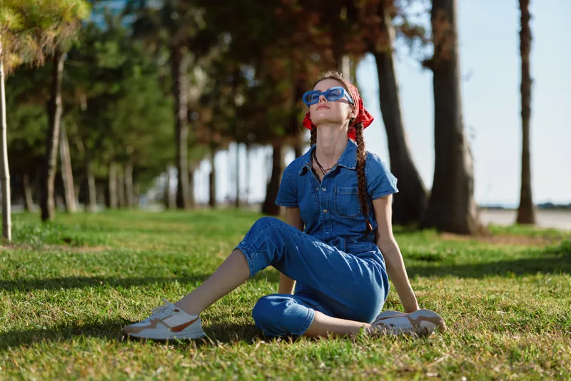 lifestyle woman in modern boho-western denim street style sitting on grass under trees with film color effects, wearing sunglasses and red bandana