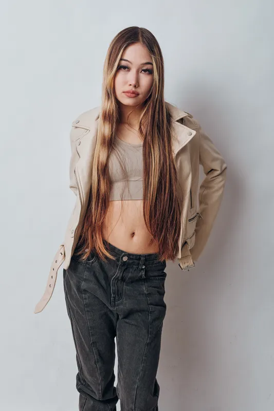 Young woman with long straight hair posing in beige jacket and cropped top against white wall, wearing black pants and looking confidently at camera in studio setting.