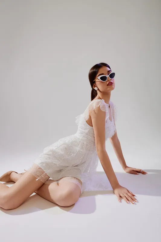 Young woman in a white lace dress and sunglasses poses confidently on the floor in a studio. Concept for beauty, fashion, and modern style.