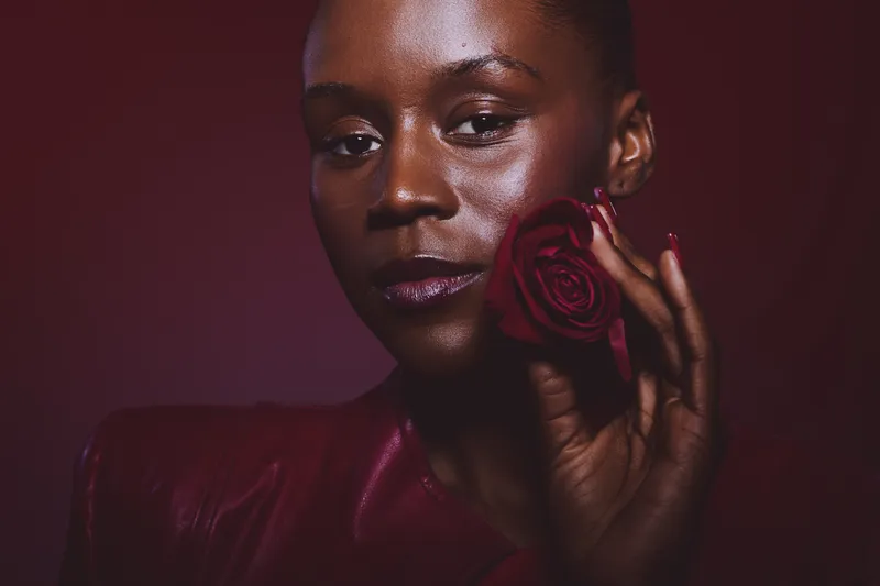 Close-up of a confident dark skinned African woman holding a red rose near her face. The deep burgundy background and lighting highlight her smooth skin and elegant expression.