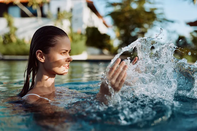 Young woman in swimwear enjoys summer day in outdoor pool splashing water with hands with blurred house and trees in background under clear sky.