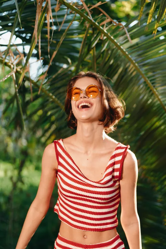 A cheerful young woman wearing a striped red and white top and orange sunglasses enjoys a sunny outdoor setting, radiating happiness and carefree summer energy.